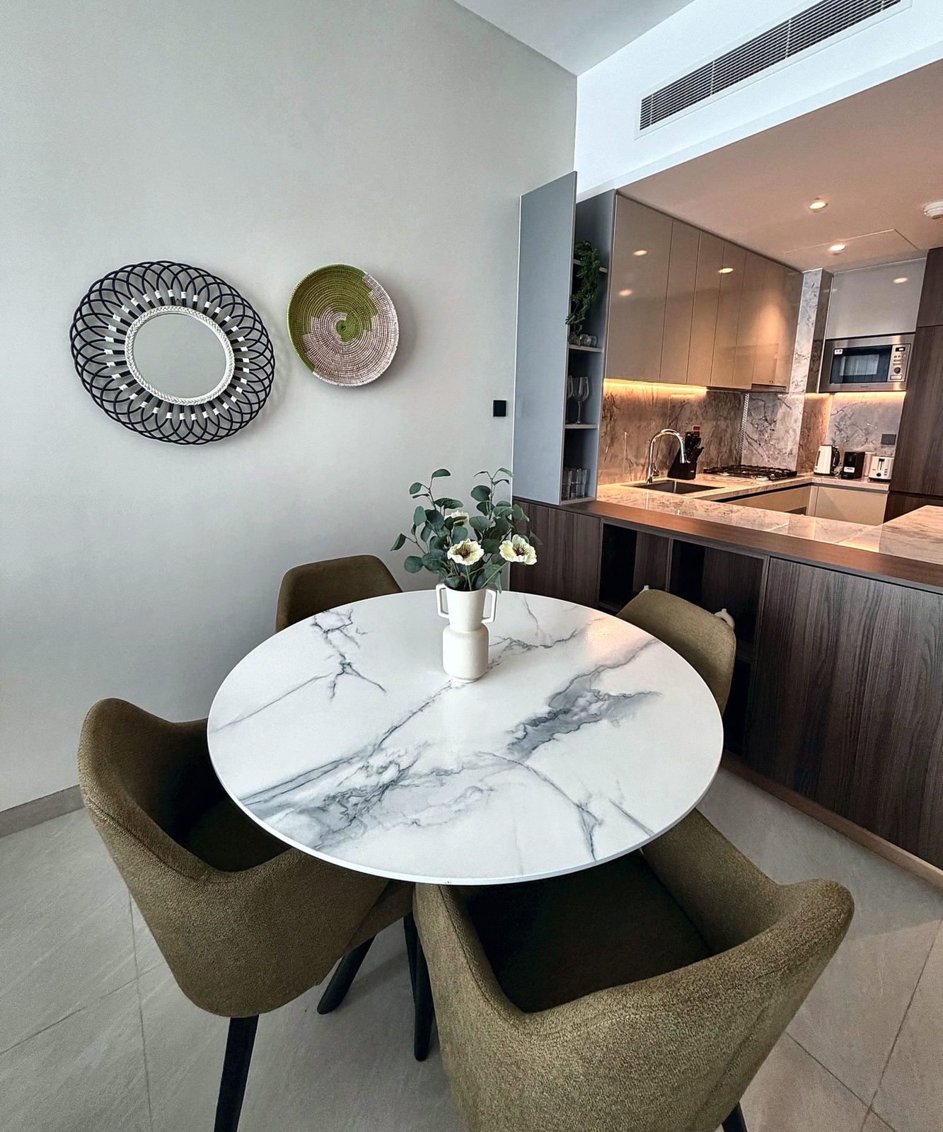 Round marble dining table with olive-green chairs, woven wall mirror and basket